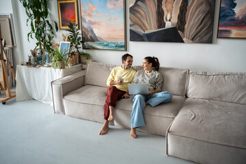 Cheerful couple spouses relaxing on couch after working day. Man with mug, woman with laptop,...