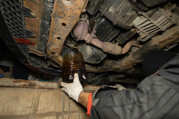 A mechanic's hands hold a canister under the bottom of a car in a pit in a garage while changing...