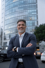 Smiling confident latin hispanic mature business man standing crossed arms on busy city street. Proud vertical portrait of older senior businessman professional entrepreneur looking camera at office
