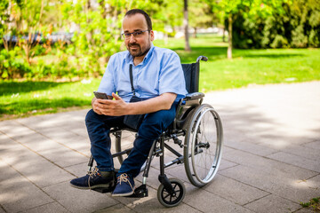 Portrait of happy man in wheelchair. He is enjoying sunny day in city park and messaging on smartphone.