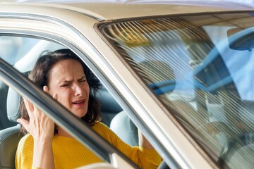 Frustrated woman in car, hand on temple, eyes closed, expression of stress, sunlit vehicle interior...