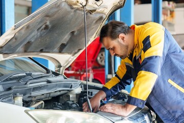 Focused mechanic in yellow and blue uniform inspects car engine, tools in hand, garage with red...