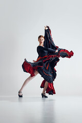Artistic capture of flamenco dancer in motion, elegant woman in black and red dress making dynamic movements over grey studio background. Concept of art of movement, classical dance, beauty, festival