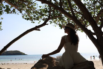 An Asian woman in a casual suit relaxes at the beach beside the sea, holding a glass of orange...