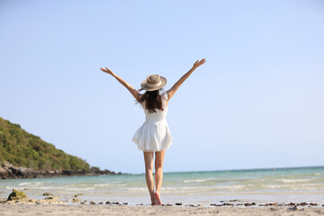 Young attractive ethnic Thai millennial woman posing at suny spreading arms to sides and various postures dressed in white casual clothes stands on the ocean with sandy beach. Love for travel