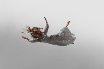 Graceful woman in diaphanous white dress drifts in space of soft light gracefully levitate against white studio background. Concept of beauty, feminine elegance and purity, dream and reality.