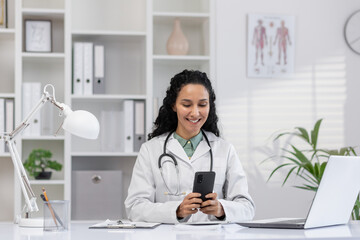 Cheerful female doctor in a white coat using a smartphone in a modern clinic office. She is smiling and appears engaged and relaxed in her professional environment.