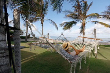 Elegant woman chilling  in a hammock strung between palm trees on the beach at a tropical resort, wearing dress and straw hat