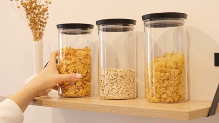 A woman carefully places a container of cereal on a kitchen shelf. The shelf already stores other containers, providing easy access and neatness. The concept of placing kitchen utensils on shelves