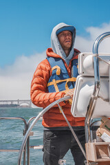 Man steering yacht in cool weather with Golden Gate Bridge in background, indicating San Francisco...