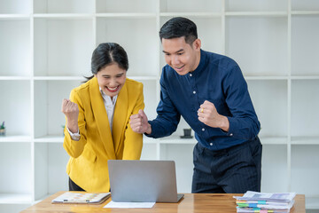 Two Asian business professionals excitedly celebrate success, looking at a laptop screen in a...