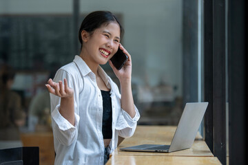 Smiling Asian young woman enjoys using her smartphone while sitting at a modern work desk with a...