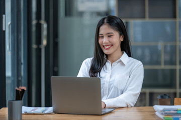 Smiling Asian young woman enjoys using her smartphone while sitting at a modern work desk with a laptop.