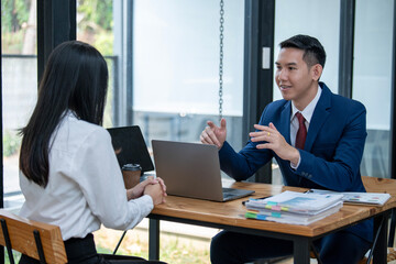 Two Asian professionals engaging in a thoughtful discussion at a business meeting, with a laptop and documents on the table.