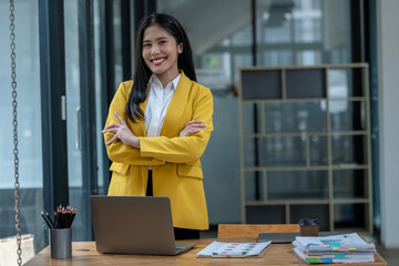 Serious Asian businesswoman in a yellow blazer stands confidently by her desk, arms crossed, in a...