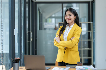 Serious Asian businesswoman in a yellow blazer stands confidently by her desk, arms crossed, in a...