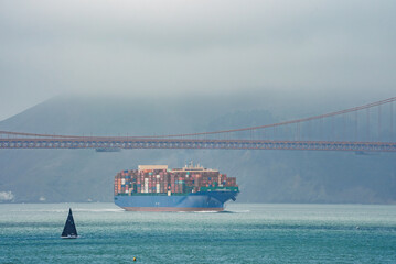 Iconic Golden Gate Bridge in San Francisco sailboat with black sail contrasts cargo ship filled...