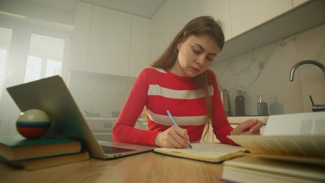A woman makes important notes in a notebook while sitting in the kitchen. She also manages to work at the laptop typing text and looking for information. High quality 4k footage