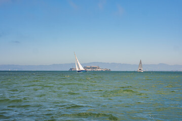 Tranquil view of San Francisco Bay with choppy waters, sailboats, Alcatraz Island silhouette, and coastal mountains in the background, capturing the bay's beauty and historical significance.