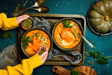 Female hands holding a bowl of pumpkin cream soup on a dark background. Autumn food concept. Top...