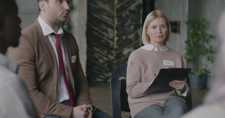 Experienced female psychologist leading therapy session with diverse group of people indoors in mental health center. Psychology and recovery concept.