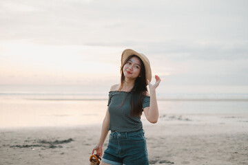 A beautiful young Asian girl is holding her straw hat with smile face expression on the beach during sunset while looking at the camera with bokeh background