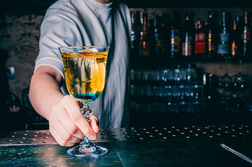 person in grey attire holds a stemmed glass containing a yellowish cocktail. The individual stands...