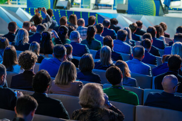 A diverse group of professionals is seen engaging at a business event, focusing intently on a speaker off-camera. The scene captures a sense of learning, networking, and professional development.