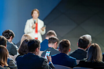 A professional woman speaks to an attentive audience at a business conference. Participants listen intently, some taking notes on digital devices.