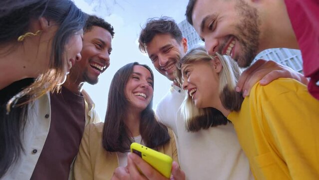 Young group of gen z people having fun using cell phone together outside. Cheerful community of student zoomers enjoying social media content on smartphone app.