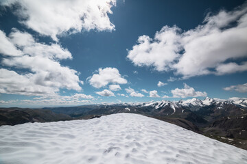 Epic vast top view from snow-capped vertex to wide alpine vastness. High mountains silhouettes with snowy tops. Snowfield at summit in sunlight. Large mountain range with snow-white peaks under clouds