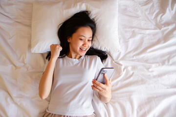 Top view of happy asian young woman awaken in white cozy bed using modern smartphone gadget, happy...