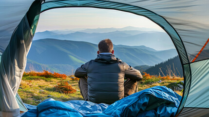 Naklejka premium Back view of a man sitting in a tent, looking out at a panoramic mountain view during sunrise.