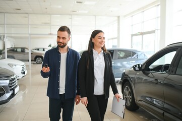 Smiling car seller standing in car salon with customer and showing around cars on sale