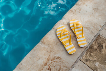 Naklejka premium Striped flipflops on the edge of an outdoor swimming pool, with blue water and beige wall in the background