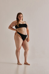 Full-length image of beautiful young girl with chubby body shape posing in black underwear against grey studio background. Concept of natural beauty, body positivity, care, acceptance