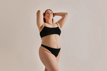 Serene expression of self-love. Young elegant girl with plus size body shape posing in underwear against grey studio background. Concept of natural beauty, body positivity, care, acceptance