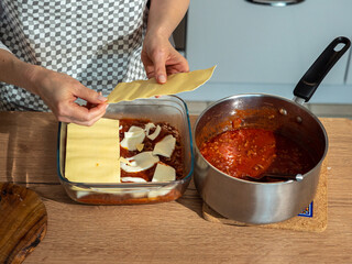 Lazzania, Italian dish, preparation, placing pasta on tomato sauce and mozzarella cheese, next to it is a metal pot with sauce, wooden countertop