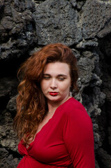 Portrait de femme rousse avec une robe rouge devant un mur en pierres noires