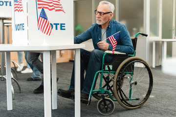 Senior man holding American flag sitting in wheelchair voting in booth with American flag
