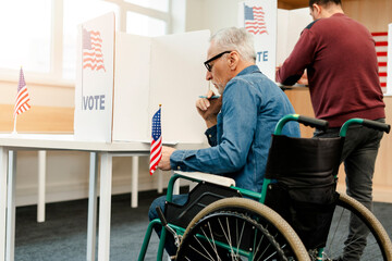 Mature, elderly man wearing eyeglasses holding American flag sitting in wheelchair voting in booth