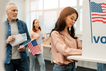 Cheerful young woman, Asian voter waiting in line, standing in polling booth with American flag