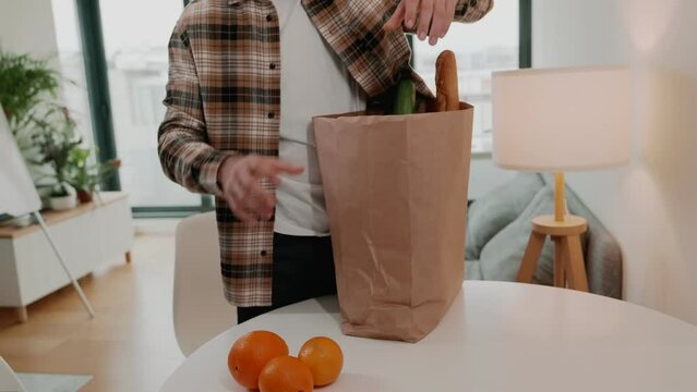 A man in a plaid shirt unpacks a paper bag full of fresh groceries on a table at home, indicating a sense of domestic life, household responsibilities, and healthy lifestyle choices.