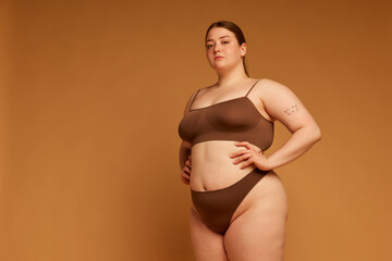 Confidence and grace. Beautiful young girl with plus size body posing in cotton underwear against light brown studio background. Concept of natural beauty, body positivity, care, acceptance
