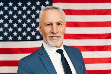 Smiling senior bearded gray haired man presidential candidate wearing suit posing on American flag