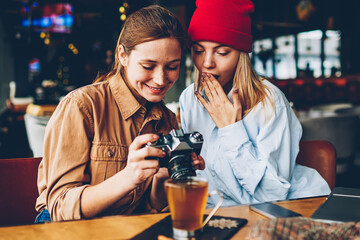 Wondered stylish teenager with cheerful friend female photographer dressed in casual outfit watching shocked photos on vintage camera during meeting in cafe interior enjoying recreation time