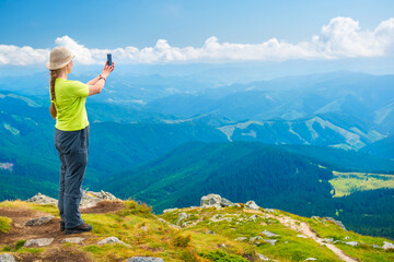 Young woman standing on mountain cliff and taking travel selfie on smartphone