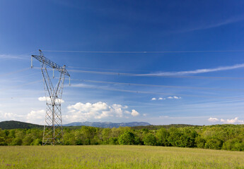 Green field against a blue sky with an electricity pylon in the foreground and hills and mountains in the background - obrazy, fototapety, plakaty