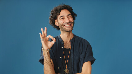 Happy attractive man with curly hair, dressed in blue shirt, shows ok gesture looking at camera...