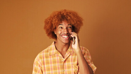 Laughing stylish curly guy speaks emotionally on mobile phone, isolated on brown background in the studio